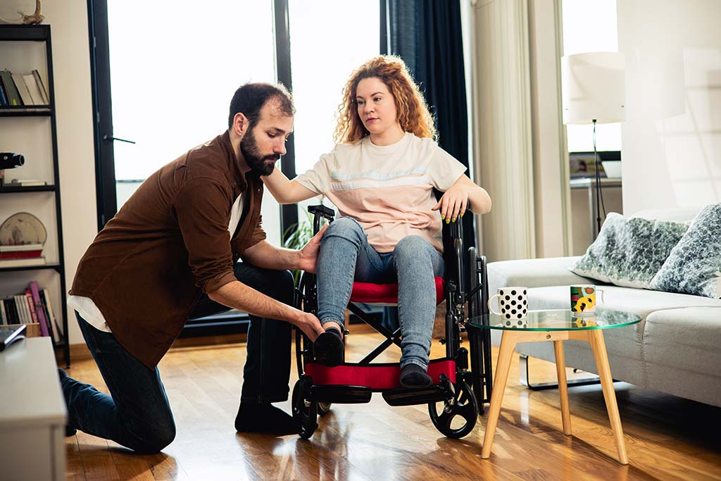 A male assistant helps a woman in a wheelchair put on a shoe in her home