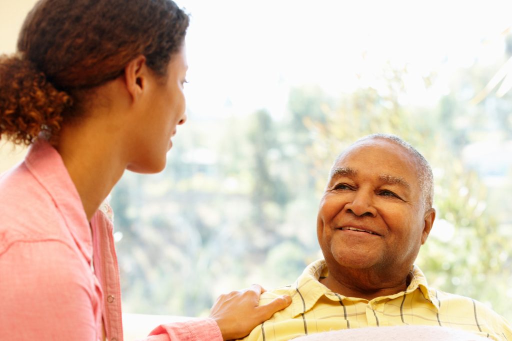 A woman chats with a seated man. She has her hand on his shoulder.