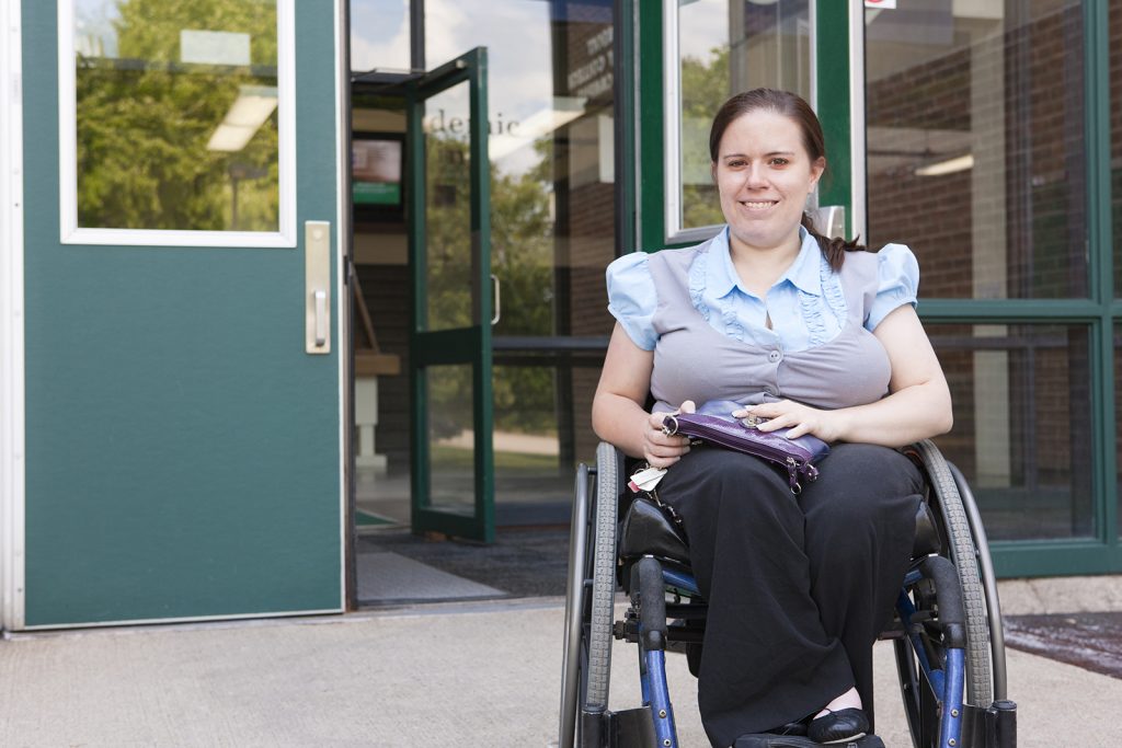A smiling woman in a wheelchair is holding a folio outside of a business building