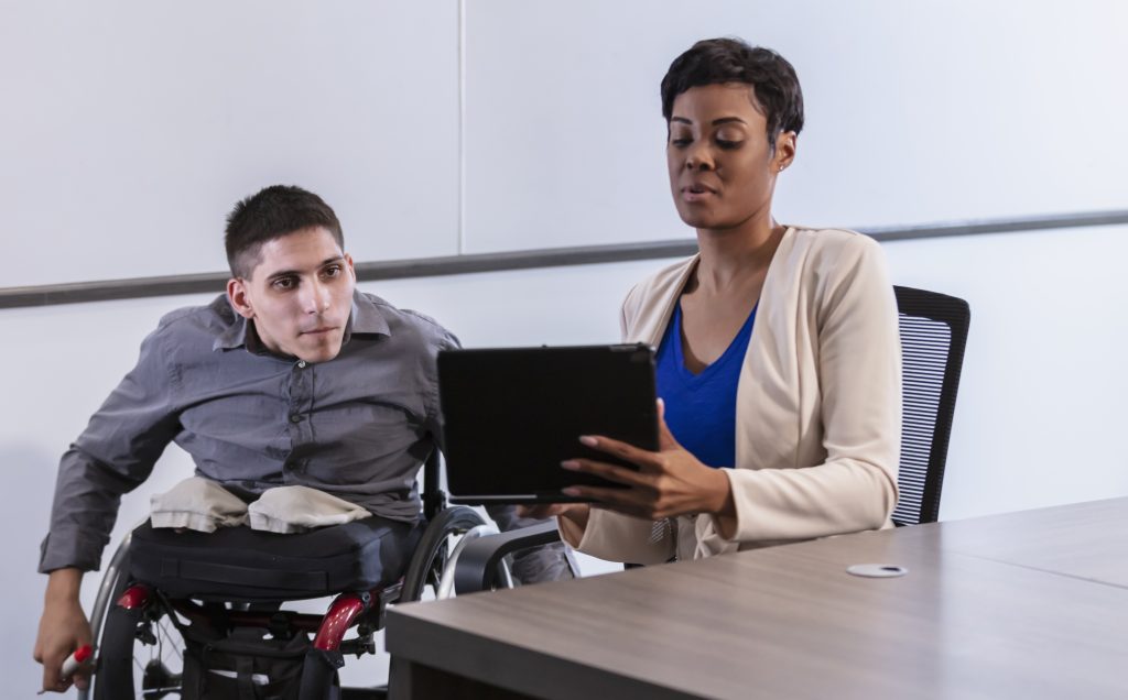 A man in a wheelchair talks with a coworker who is looking at her laptop