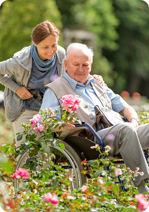 a caretaker pushing an older man in a wheelchair looking at flowers