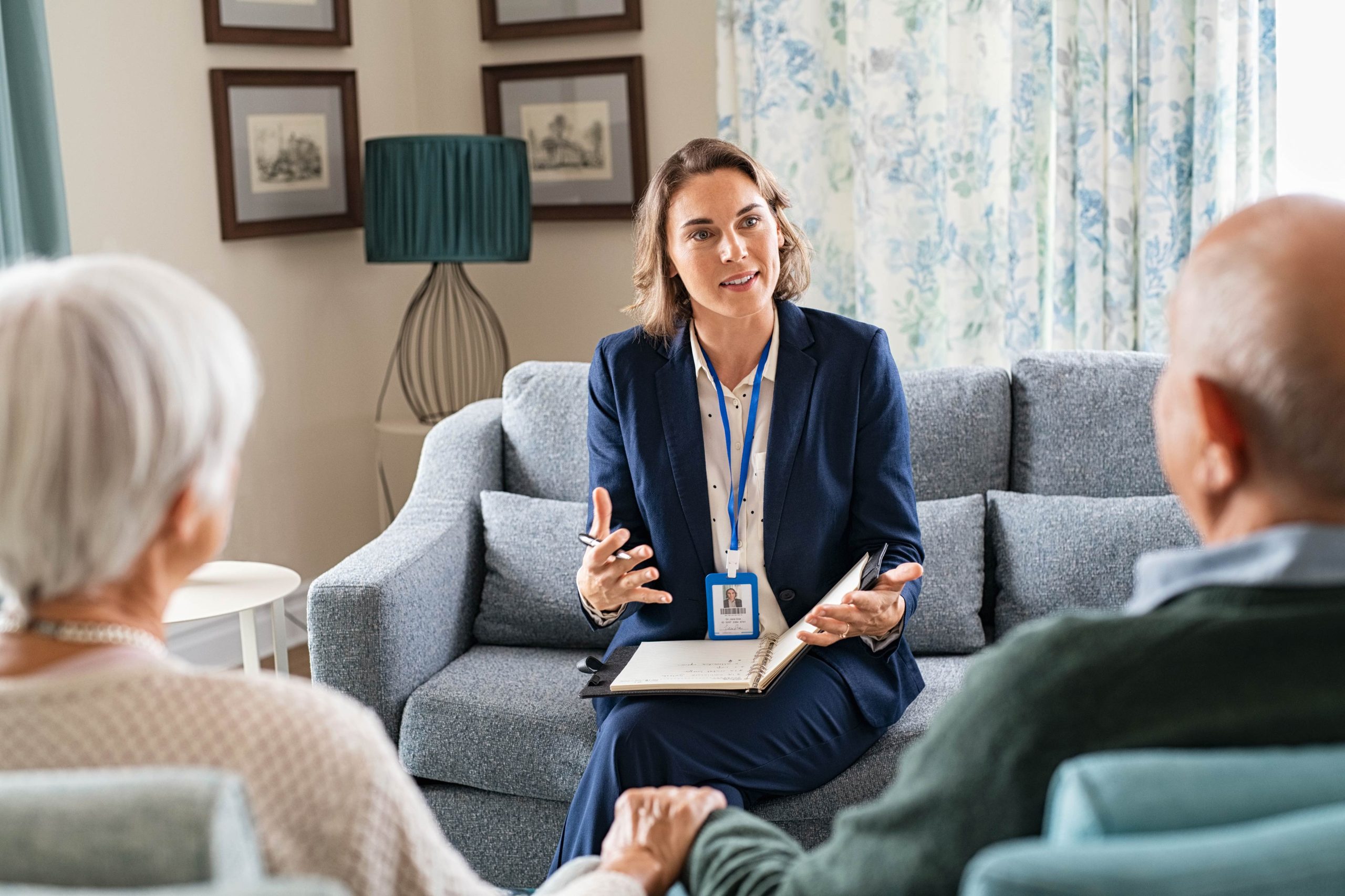 A woman in a business suit and name badge sits on a couch talking to an elderly couple in their home
