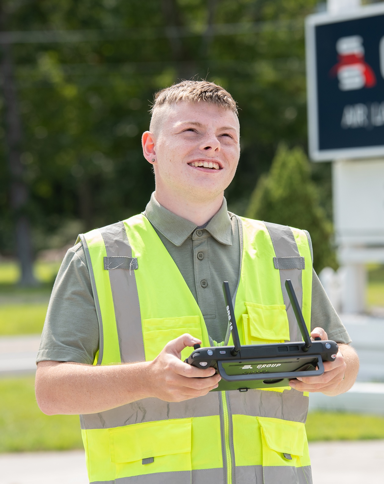 A young man wearing a safety vest uses a drone remote control while looking towards the sky