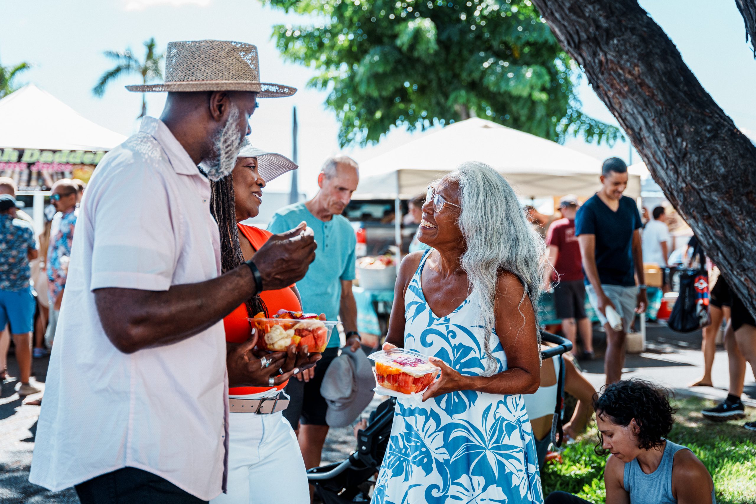 An African American senior couple eat fresh fruit and cheerfully chat with their local tour guide, a senior woman of Hawaiian decent, while at a farmer's market