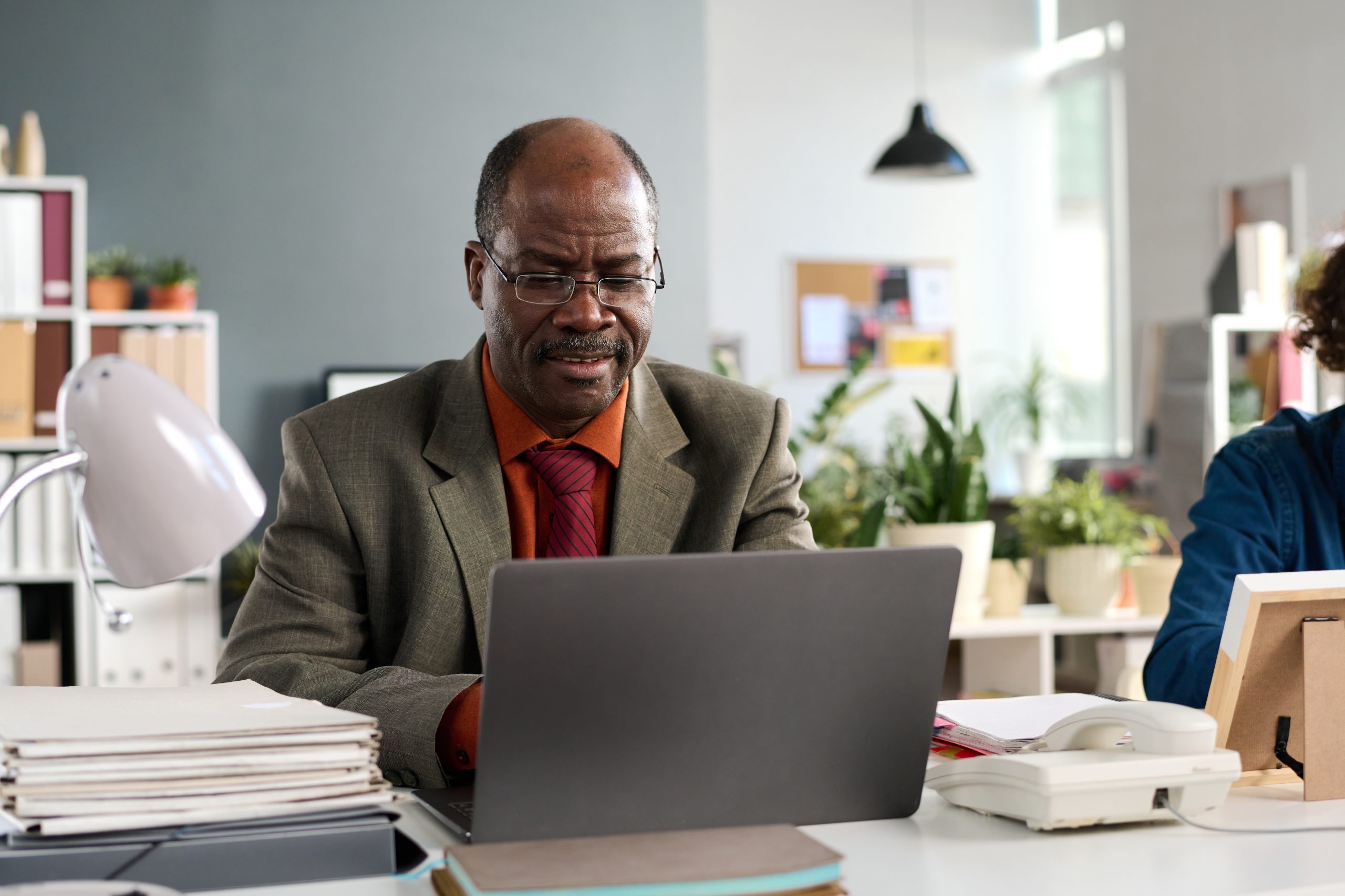 Black senior man using a laptop next to a colleague in an business office