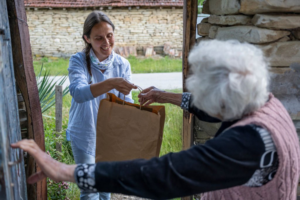 An elderly woman opens the door for a woman who is handing her a paper bag filled with food.